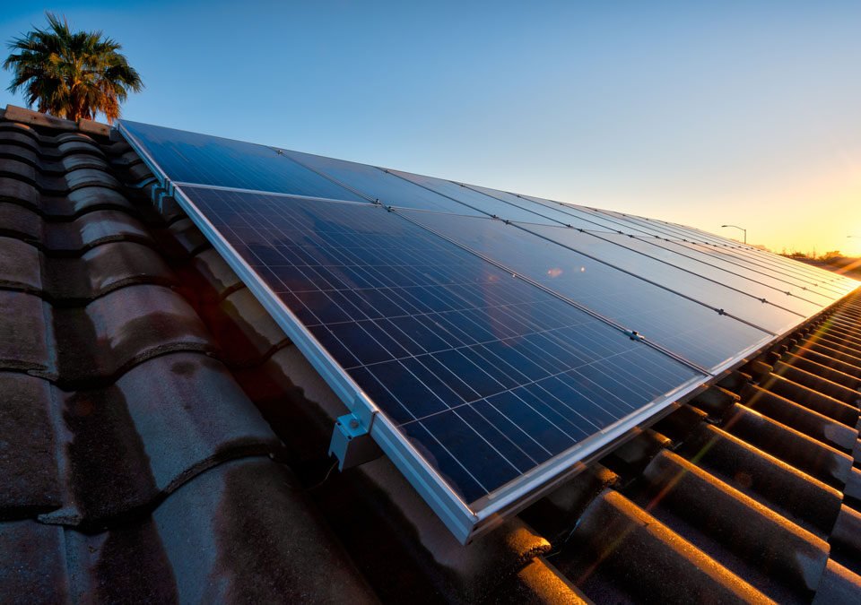 Panel solar en primer plano sobre un techo de tejas de barro naranjadas atrás se ve la copa de una palmera y en el fondo se ve un atardecer con un cielo azul