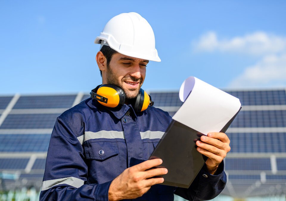 Trabajador en primer plano revisando planilla en el fondo se ven paneles solares y un cielo azul