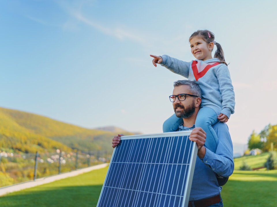 Un padre con su hija sobre sus hombros mirando hacia el futuro, sostiene en la mano un panel solar y en el fondo se ve un campo verde y cielo azul