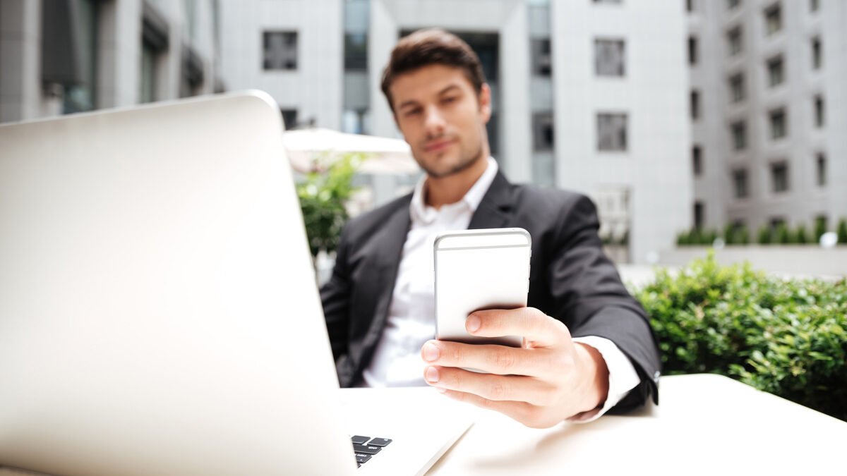 Hombre con traje de negocios revisando su celular y al frente de su laptop, al fonde se ven edificios de oficinas