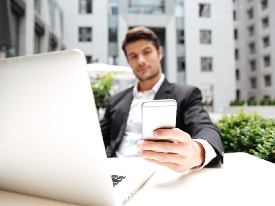 Hombre con traje de negocios revisando su celular y al frente de su laptop, al fonde se ven edificios de oficinas
